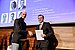 Two men shake hands on a stage and smile into the camera. The one on the right holds a certificate in his hand.