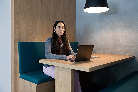 ine Studentin sitzt an einem Tisch mit Laptop und lacht in die Kamera.