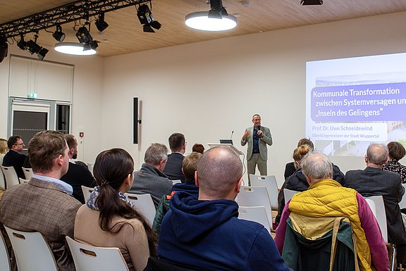 Foto aus dem Publikum einer Veranstaltung, ein Redner steht vorne und spricht über den kommunalen Wandel.