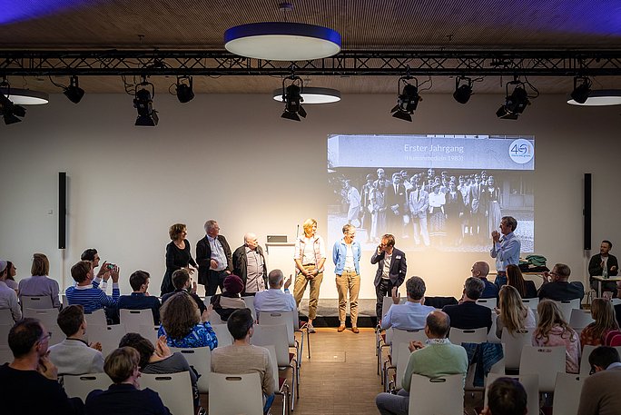 Foto eines Veranstaltungssaals: Eine Gruppe steht im Scheinwerferlicht vor dem Publikum. Im Hintergrund wird an Schwarzweiß-Foto an die Wand projiziert.