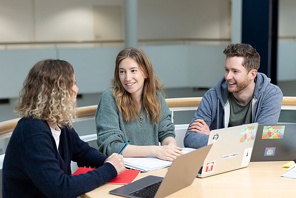 Drei junge Menschen sitzen an einem Tisch und unterhalten sich. Vor ihnen stehen drei Laptops.