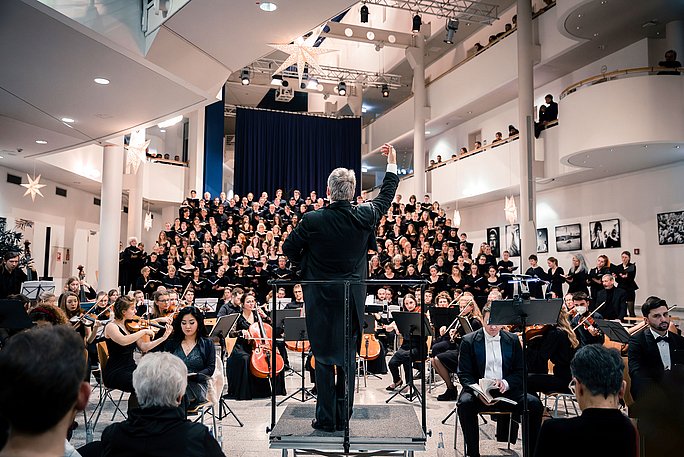 Im Vordergrund der Dirigent mit Blick Richtung Orchester und Chor. Alle Musiker:innen sind schwarz gekleidet. Die Haupthalle der Uni ist mit weihnachtlichen Sternen geschmückt.