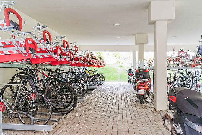 Blick in die Fahrradgarage der Uni. Hier stehen Fahrräder in Metallständern auf zwei Ebenen. Auf der rechten Bildseite stehen außerdem zwei Motorroller.