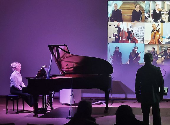 Eine Frau sitzt an einem Piano, auf einer Wand hinter ihr sieht man Musiker:innen, die digital zugeschaltet sind.