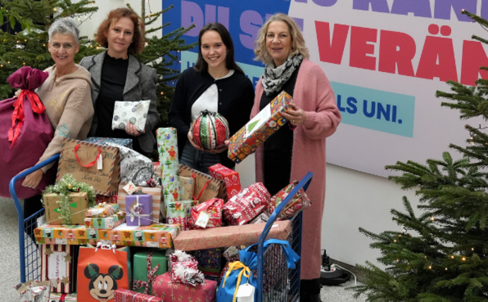 Four women are standing between Christmas trees, holding Christmas presents in their hands. In front of them is a trolley with more parcels.