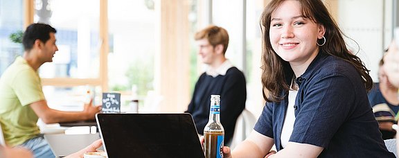 Eine junge Frau sitzt mit einem Softdrink an einem Tisch vor einem Laptop.