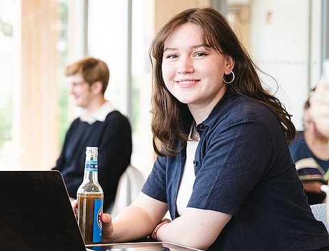 Eine junge Frau sitzt mit einem Softdrink an einem Tisch vor einem Laptop.