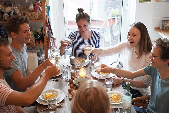 Eine Gruppe Studierender sitzt gemeinsam beim Abendessen am Esstisch und stößt mit Getränken an.