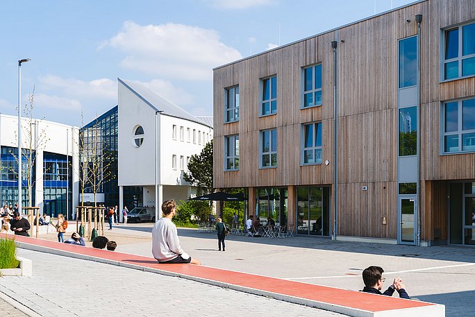 Übersicht des Campus der Uni Witten/Herdecke. Links ist das weiß-blaue Hauptgebäude, rechts das Holzgebäude. Im Vordergrund sitzen Menschen auf Betonsstufen.