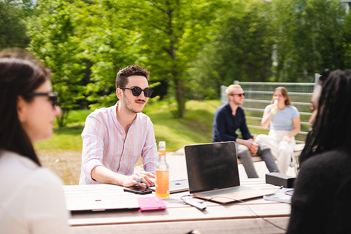 Eine Gruppe Studierender sitzt gemeinsam an einem Tisch auf dem Campus. Da es sonnig und warm ist, tragen sie Sonnenbrillen und sommerliche Kleidung. Zwischen ihnen stehen ein Laptop, Lernunterlagen und Getränke.