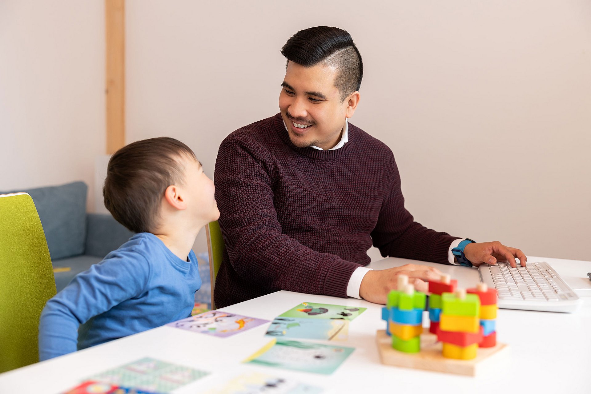 Ein Vater sitzt gemeinsam mit seinem Sohn im Eltern-Kind-Raum der Uni. Vor ihnen auf dem Tisch liegen neben der Tastatur des Arbeitsrechners verschiedene Spielsachen ausgebreitet.