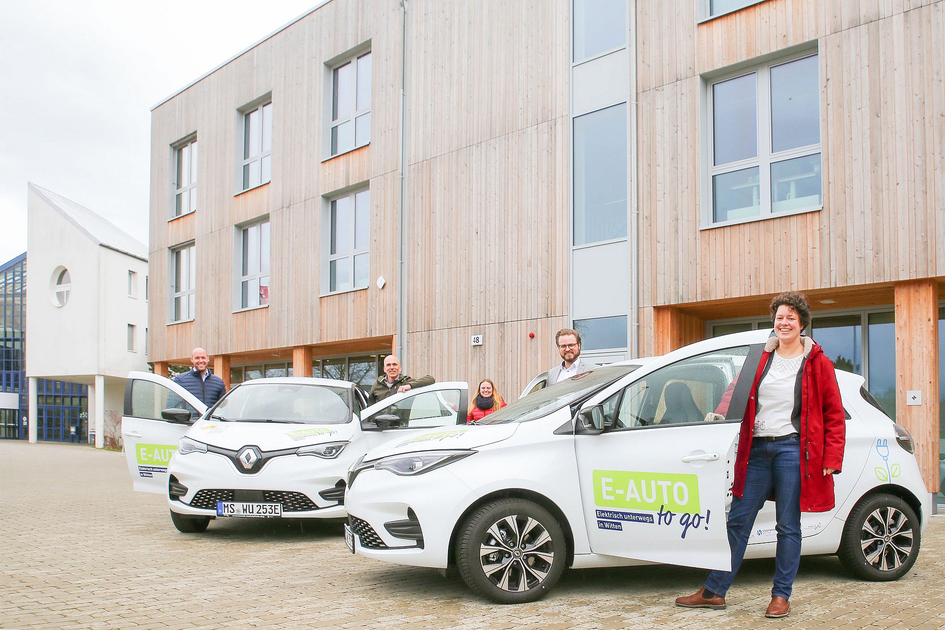 Vor dem Neubau stehen zwei weiße E-Autos mit geöffneten Fahrertüren. Daneben stehen die projektverantwortlichen Personen.