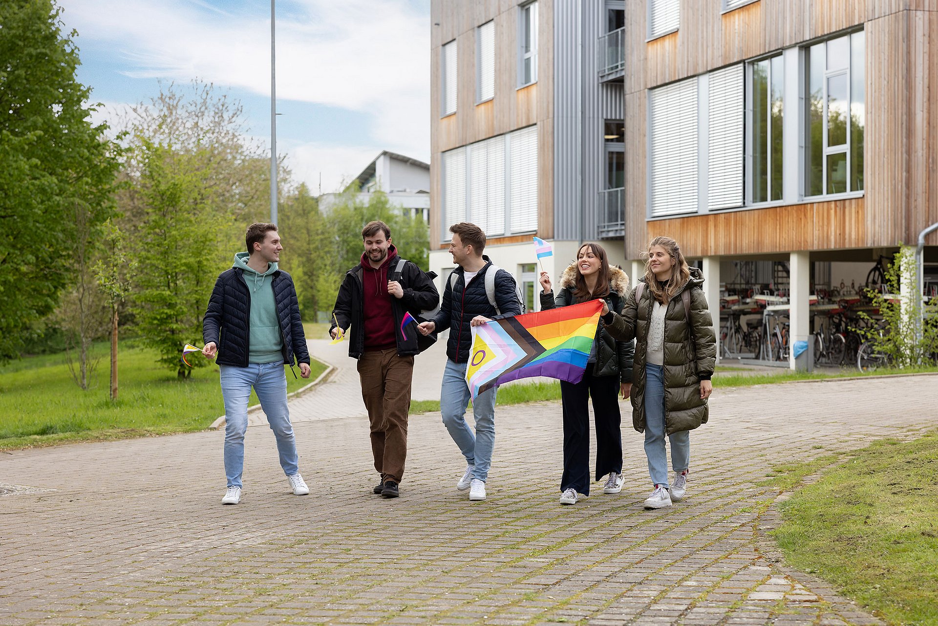 Eine Gruppe Studierender läuft mit geschwenkter LGBTQIA-Flagge über den Campus. Hier ihnen ist ein Holzgebäude.