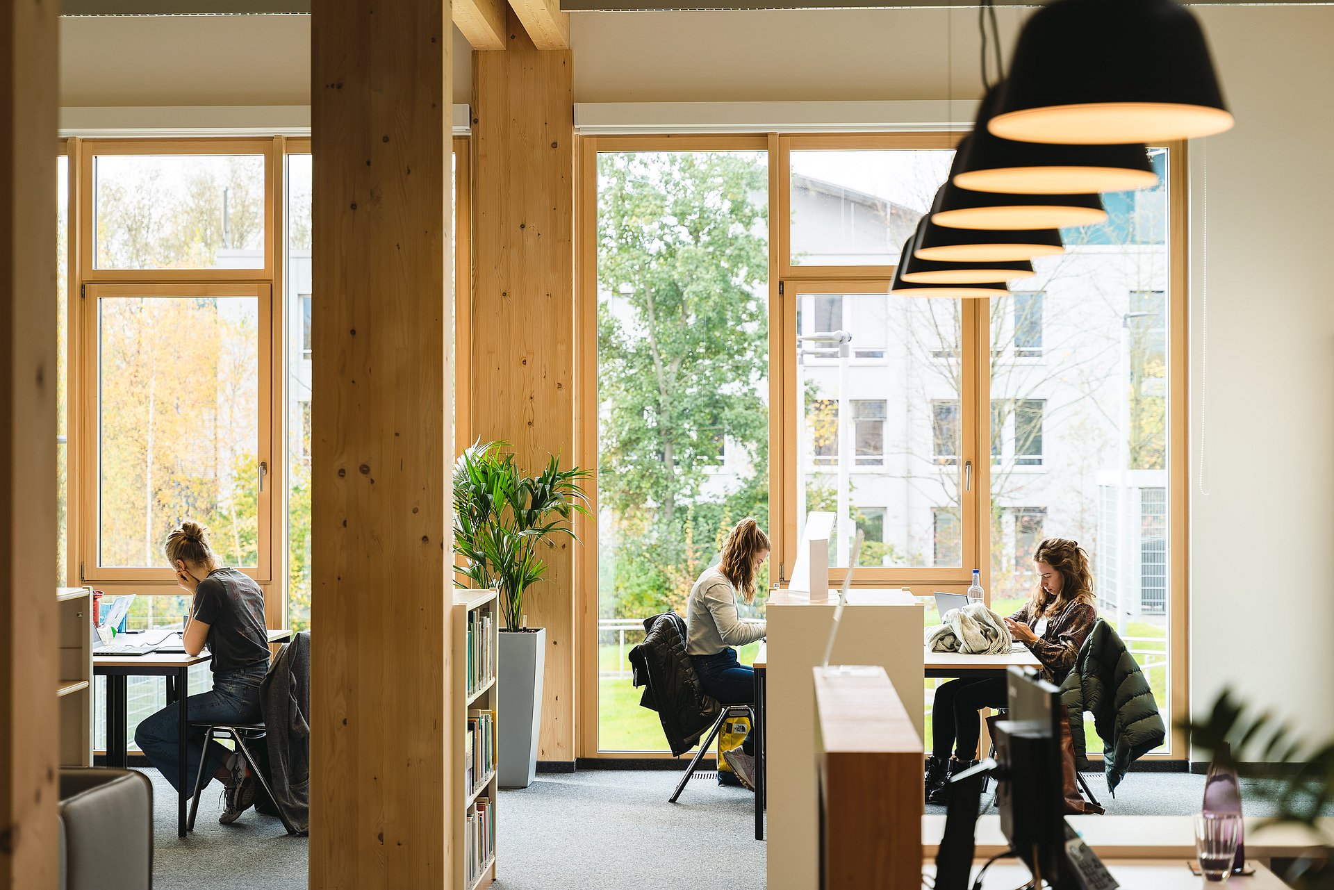 In einer ruhige Bibliothek mit großen Fenstern sitzen drei Personen an Tischen und arbeiten konzentriert an Laptops oder schauen auf ihr Smartphone.