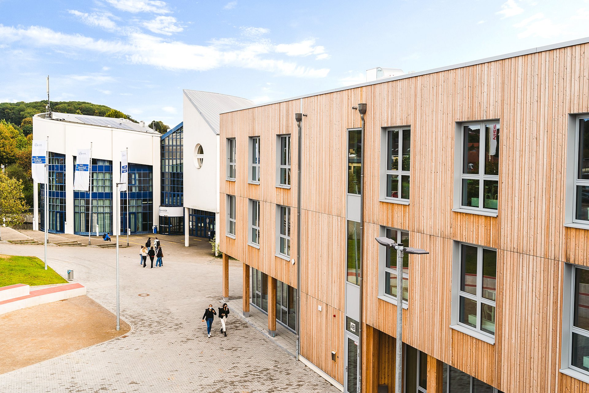 Moderne Universitätsgebäude mit Holz- und Glasfassaden sind von einem gepflasterten Innenhof umgeben. Einige Menschen gehen über den Platz, im Hintergrund ist eine grüne Landschaft zu sehen.