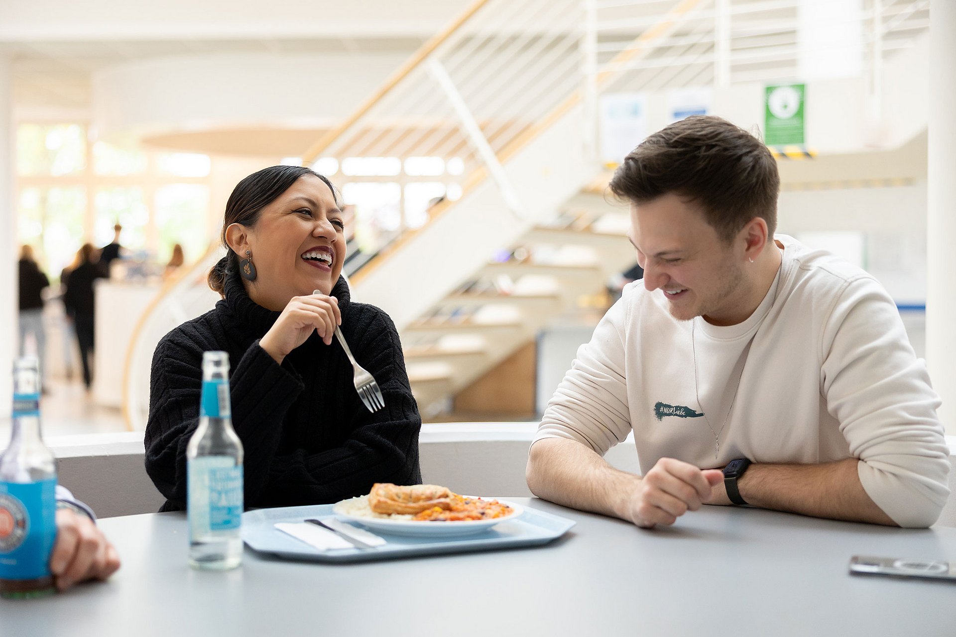 Eine Studentin und ein Student sitzen nebeneinander an einem Cafeteria-Tisch. Sie unterhalten sich und lachen. Zwischen ihnen steht ein Teller mit Essen.