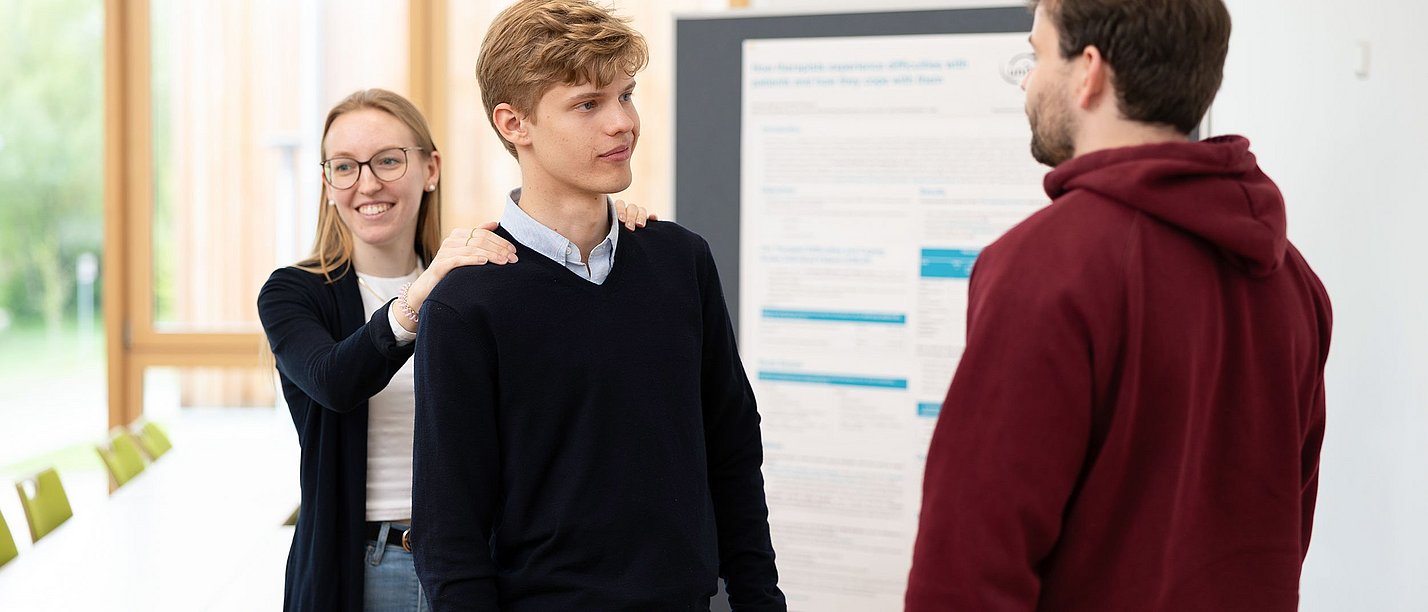 A student stands with his back to a female student and looks at another student. The student has placed her hands on his shoulders.