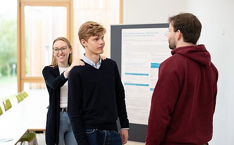 A student stands with his back to a female student and looks at another student. The student has placed her hands on his shoulders.