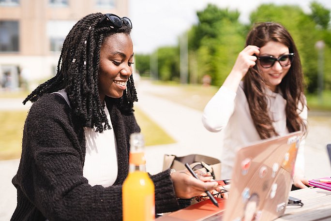 Zwei Studentinnen lernen mit Laptop auf dem Campus und bei Sonnenschein.