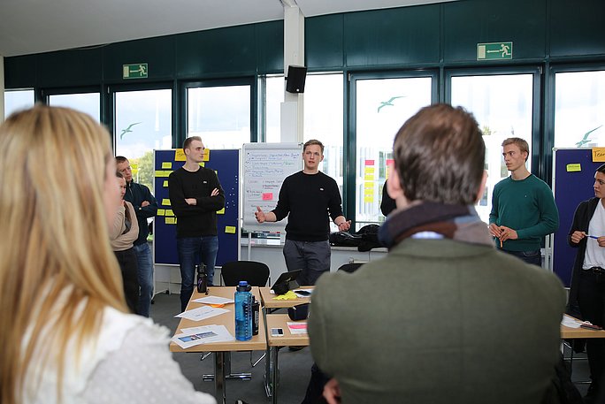 Studierende stehen vor Pinnwänden in einem Seminarraum und präsentieren ihre Ideen. 