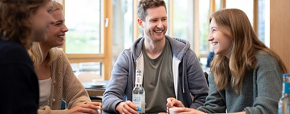 Eine Gruppe Studierender trinkt gemeinsam Kaffee. Vor ihnen stehen Getränke und Gebäck.