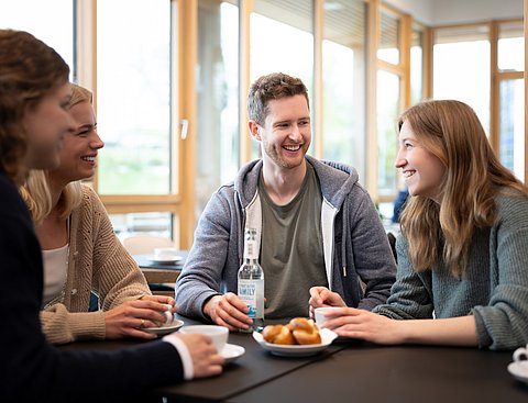 Eine Gruppe Studierender trinkt gemeinsam Kaffee. Vor ihnen stehen Getränke und Gebäck.