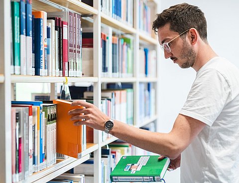 Ein Student steht vor einem Bücherregal und greift nach einem Buch.