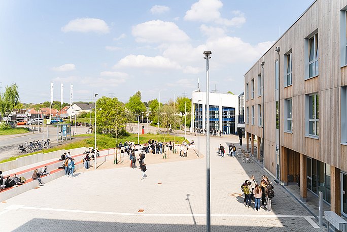 Blick über den UW/H-Campus. Rechts ist das Holz- und im Hintergrund das Hauptgebäude zu sehen. Auf dem Vorplatz laufen, stehen oder sitzen Studierende.