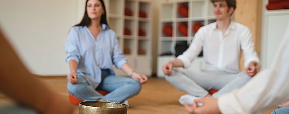 Im Fokus sind zwei Studierende, die im Schneidersitz auf dem Boden sitzen. Zwischen ihnen steht eine Klangschale.