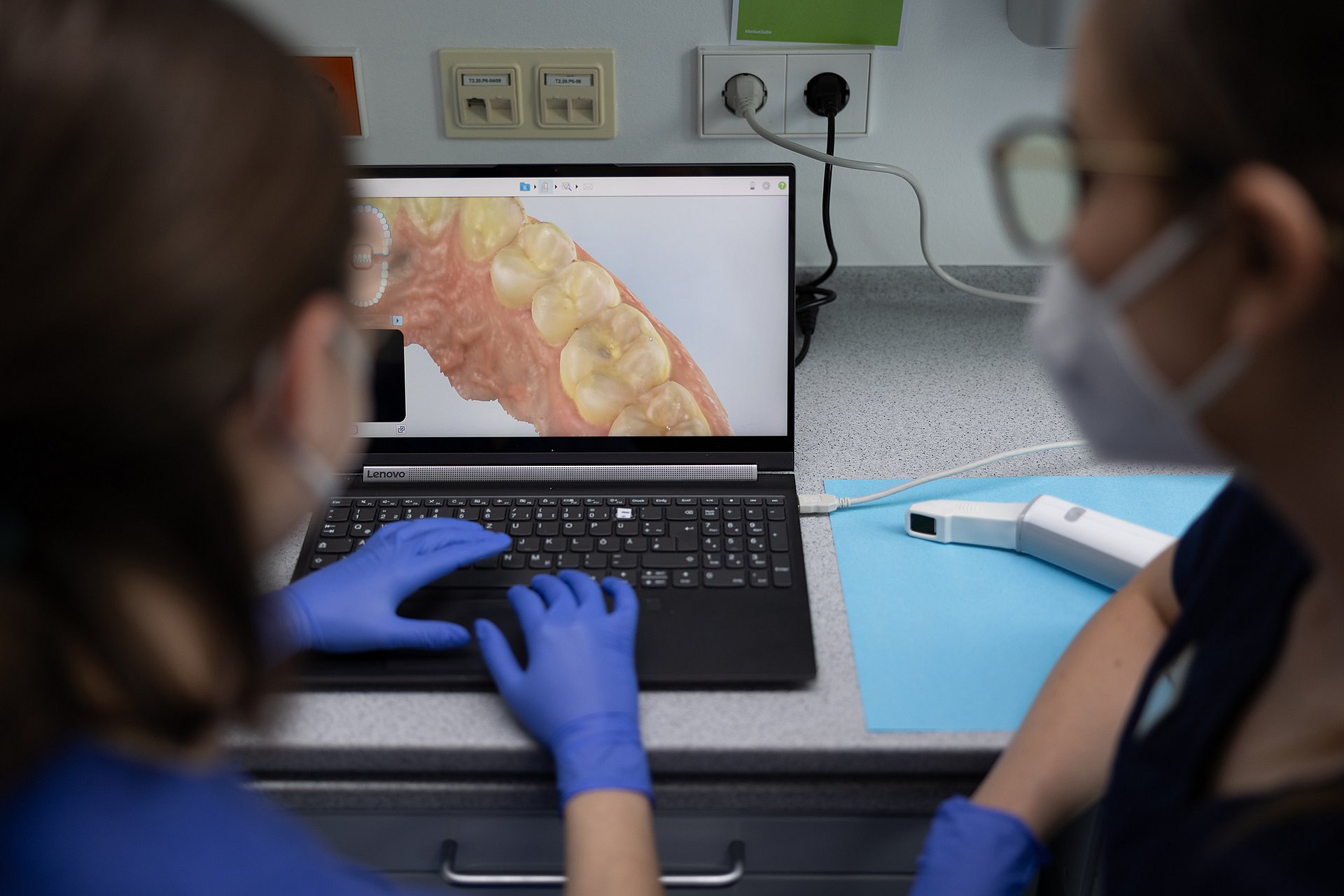 Das Bild zeigt einen Blick über die Schulter von zwei Studentinnen der Zahnmedizin. Sie tragen medizinische Handschuhe, einen Mundschutz und blaue Arbeitskleidung. Beide Frauen schauen auf einen Laptop-Bildschirm, auf dem der Ausschnitt eines Gebisses zu sehen ist.