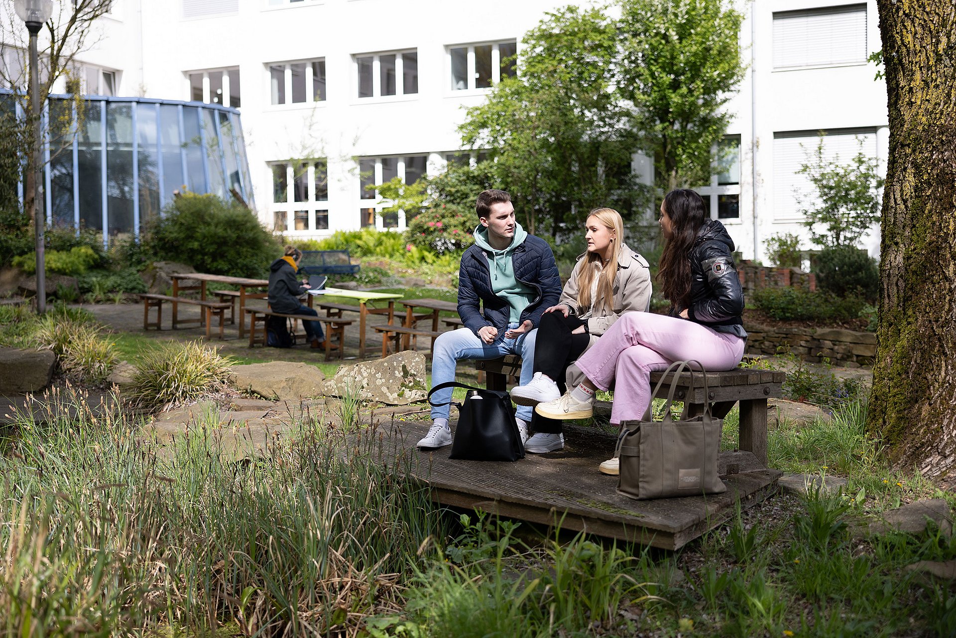 Drei Studierende sitzen auf einer Bank im grünen Unigarten. Hinter ihnen ist das Unigebäude zu erkennen.