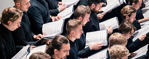 Nahaufnahme eines Uni-Chors. Alle Sänger:innen halten Notenblätter und Liedtexte in den Händen.