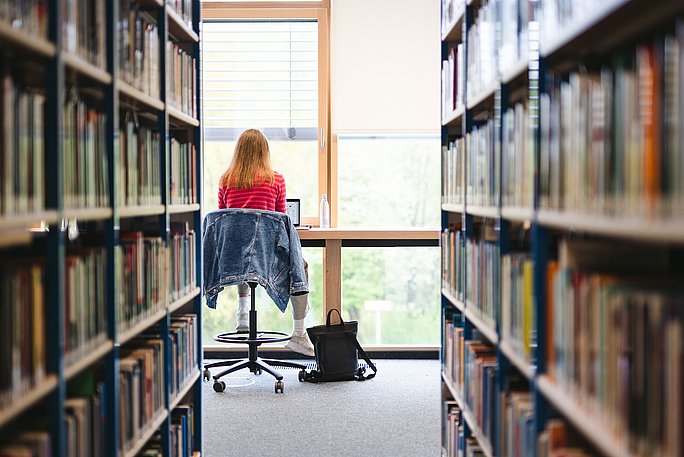 Blick zwischen zwei Bücherregalreihen. Am Ende des Ganges sitzt eine Studentin an einem Thekentisch am Fenster und lernt.