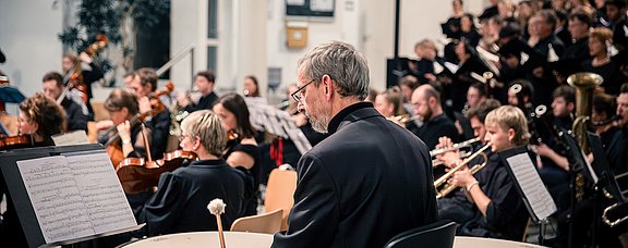 Im Fokus ist ein Paukenspieler. Im Hintergrund sitzt das restliche Orchester mit Streichinstrumenten.