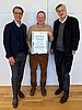 Three men are standing in front of a white man, the man in the middle is holding a certificate.