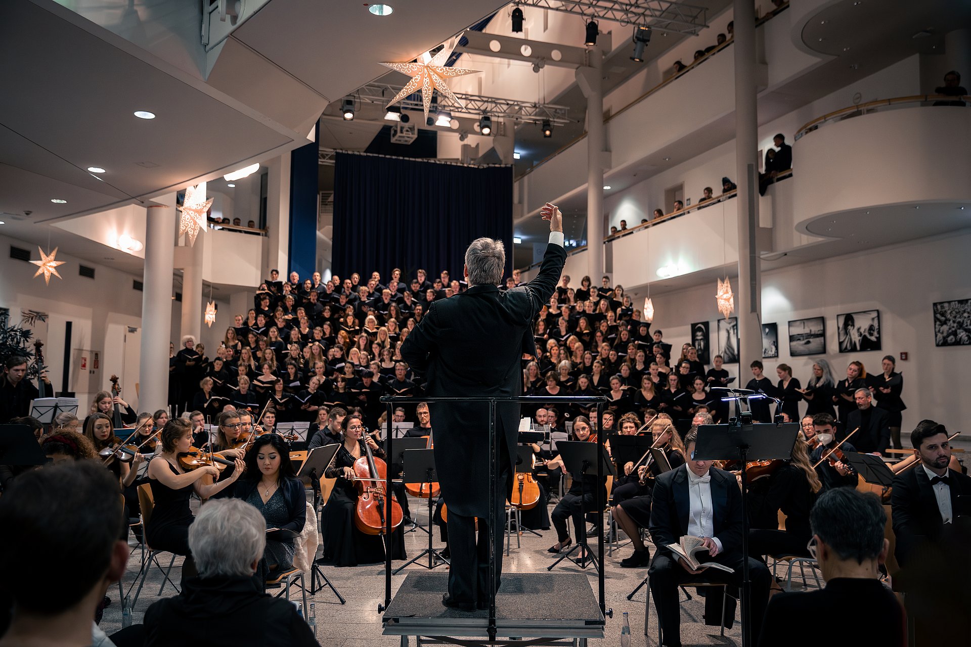 Ein Dirigent steht mit dem Rücken zum Betrachter und dirigiert einen Chor und ein Orchester.