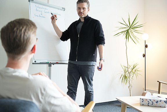 Ein Therapeut und ein Patient in einer Gesprächssituation, der Therapeut schreibt Ergebnisse auf ein Flipchart.