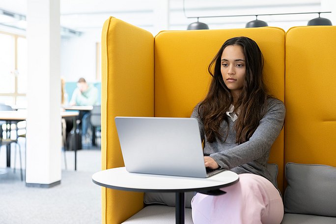 Eine Studentin sitzt in einer gelben Sitzecke in der Bibliothek. Vor ihr steht ihr Laptop auf einem kleinen runden Tisch.