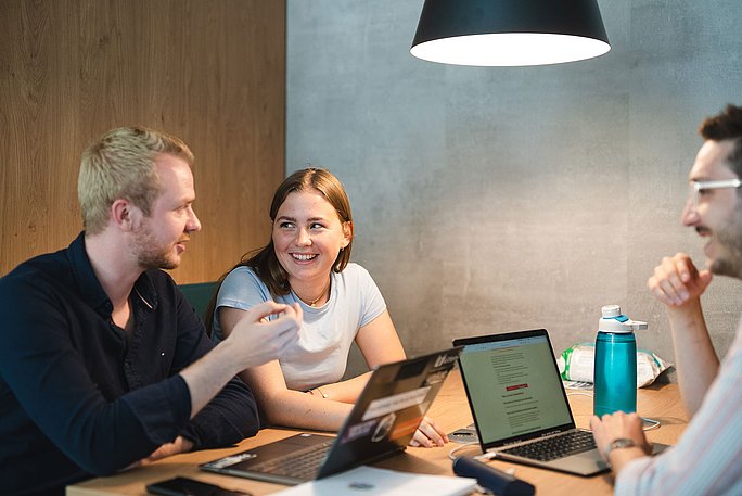 Drei Studierende sitzen gemeinsam an einem Tisch und unterhalten sich. Zwischen ihnen stehen Laptops zum Lernen.