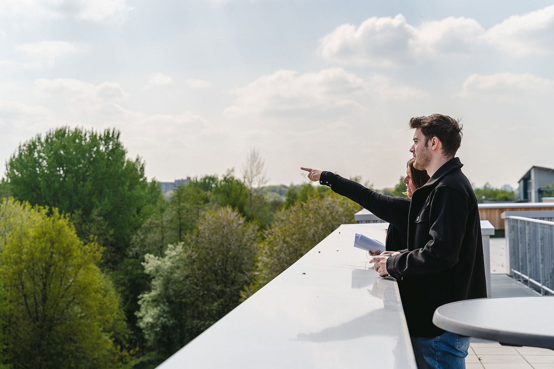 Zwei Personen stehen auf einer Dachterrasse und schauen ins Grüne. Die hintere Person deutet mit dem Finger in die Ferne.