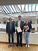 Three people smile frontally into the camera. The man in the middle holds a certificate into the picture.