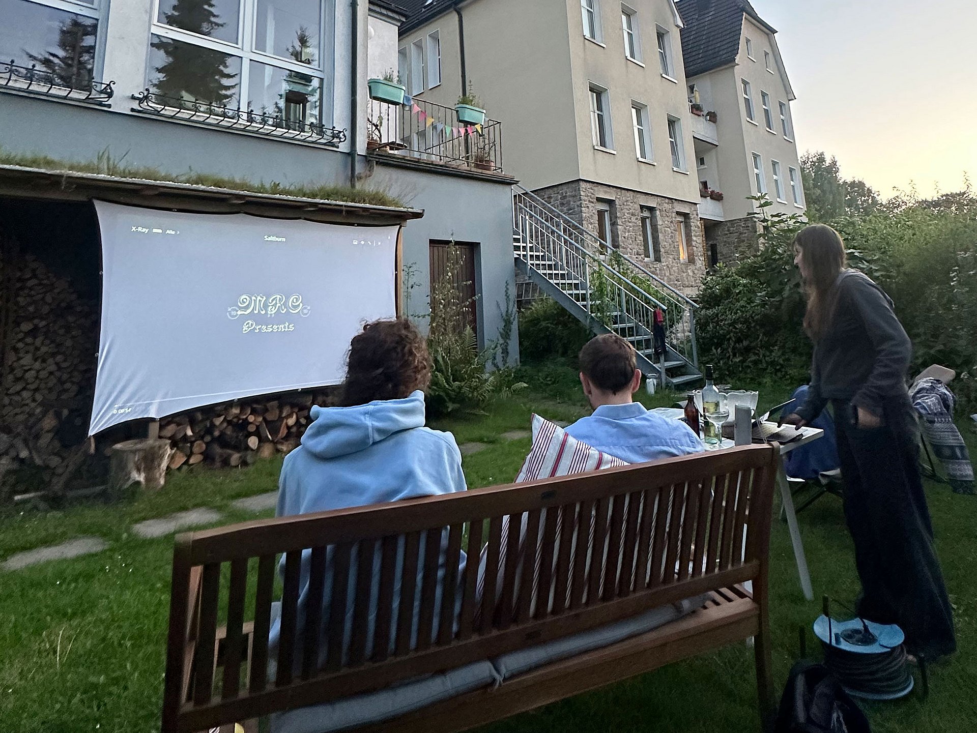 Eine Gruppe junger Menschen sitzt auf Bänken und Gartenstühlen auf einer Wiese. Sie schauen auf eine Leinwand, auf die ein Film projiziert wird. Im Hintergrund sind Häuserfassaden.