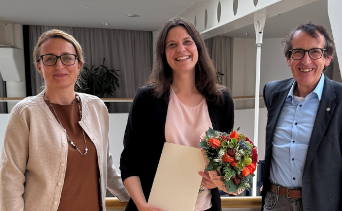 Drei Personen stehen im Hauptgebäude der Uni Witten/Herdecke. Die Person in der Mitte ist Prof. Dr. Anke Haberkamp. Sie hält ihre Berufungsurkunde in der Hand und lächelt freundlich in die Kamera.