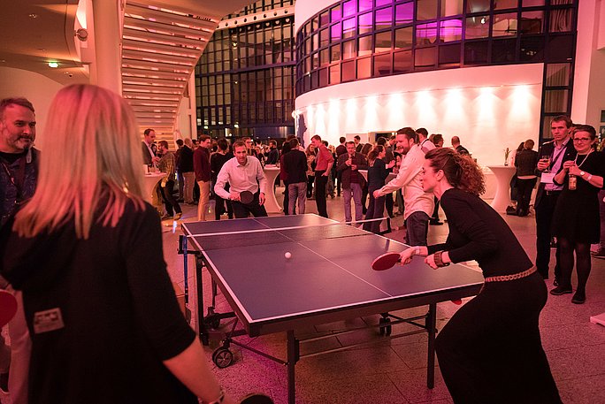 Zwei Personen spielen Tischtennis in einem Veranstaltungsfoyer. Im Hintergrund stehen weitere Personen, die zuschauen oder sich unterhalten.