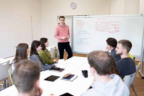 Ein junger Mann steht an einem Whiteboard und schaut in die Gesichter junger Menschen, die im Kreis an einem Tisch vor ihm sitzen.