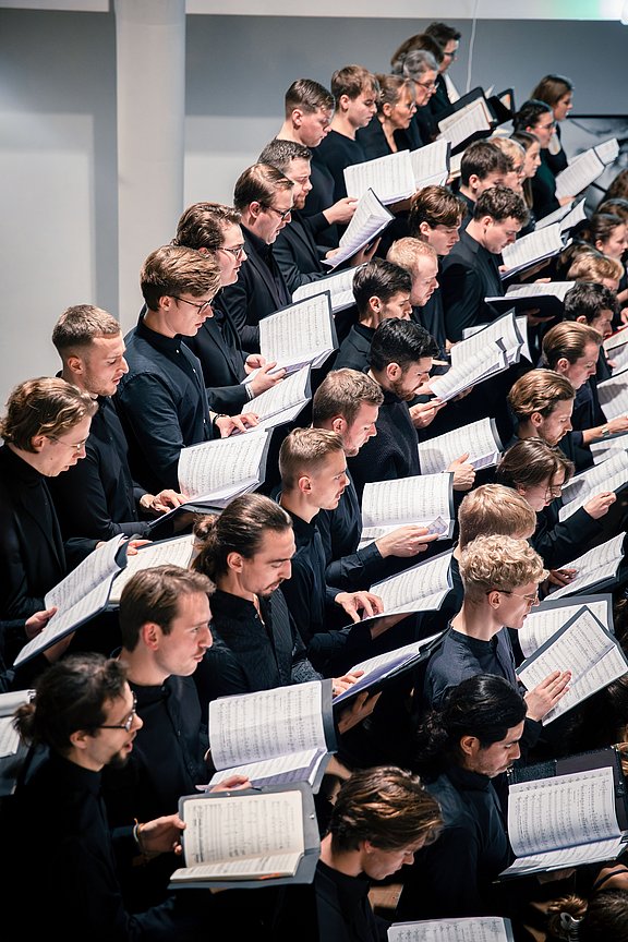 Foto eines Chors mit mehreren Sänger:innen, die Notenblätter in den Händen halten.