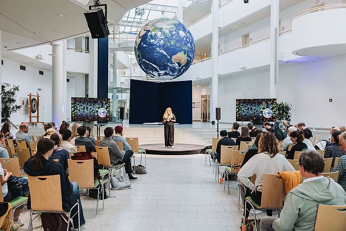 In der Haupthalle der UW/H sitzen Konferenzteilnehmende und blicken zur Rednerin in der Mitte der Halle. Von der Decke hängt eine große Erdkugel.