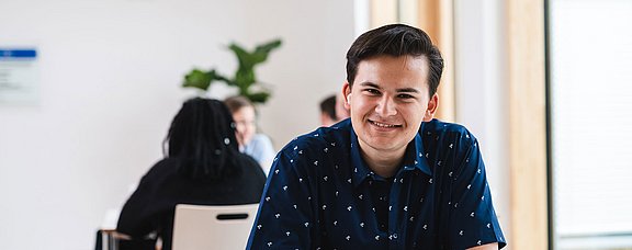 Ein junger Mann sitzt an einem Tisch und hat ein Heft vor sich liegen.