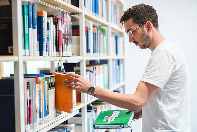 Ein Student steht vor einem Bücherregal und greift nach einem Buch.