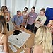 A group of people are standing in a circle around a table. They are looking together at a poster lying on the table.